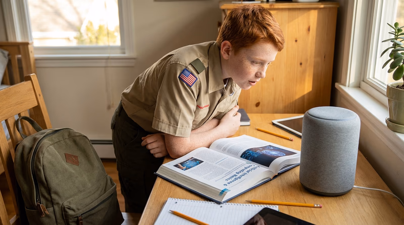 A Scout in their bedroom talking to a smart speaker (voice assistant) on their desk, with homework spread out and a backpack nearby. Natural afternoon light from a window.