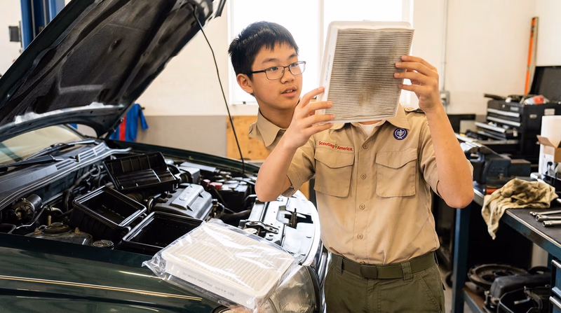 A Scout holding up a used engine air filter to the light to check its condition, with a new clean filter sitting on the fender for comparison