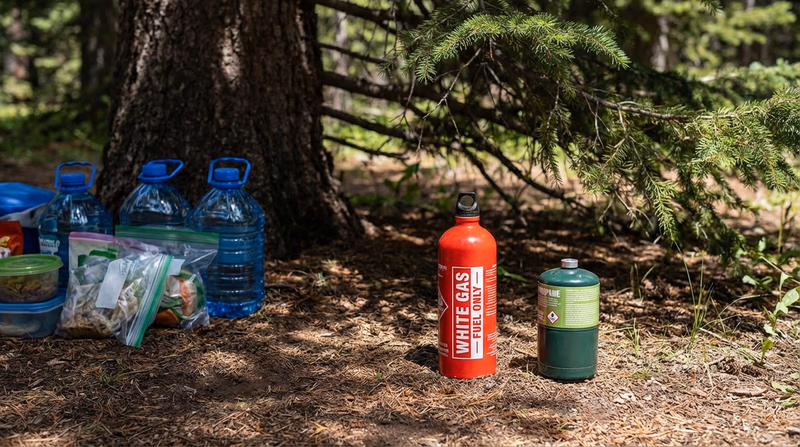 A labeled fuel bottle and a propane canister stored upright in a shaded area at camp, clearly separated from food and water supplies