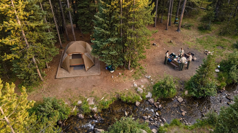 An illustrated birds-eye view of an ideal campsite showing the tent on level ground, 200 feet from a stream, behind a natural tree windbreak, with the cooking area downwind and a bear bag hung away from the sleeping area