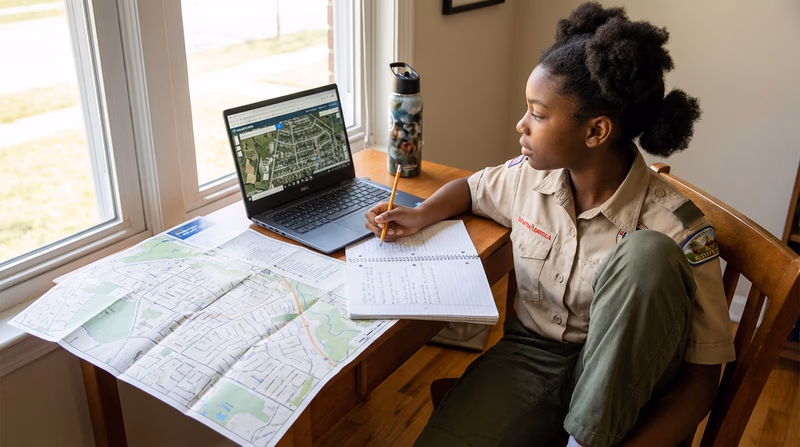 A Scout sitting at a desk using a laptop to explore a digital map, with a printed community map and notebook beside them