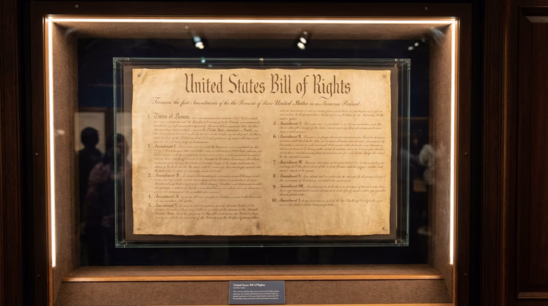 The Bill of Rights document displayed in a museum case with dramatic lighting, showing the handwritten text of the first ten amendments