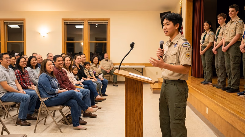 A Scout in uniform standing at a podium during a court of honor ceremony, with award recipients lined up and families watching from chairs