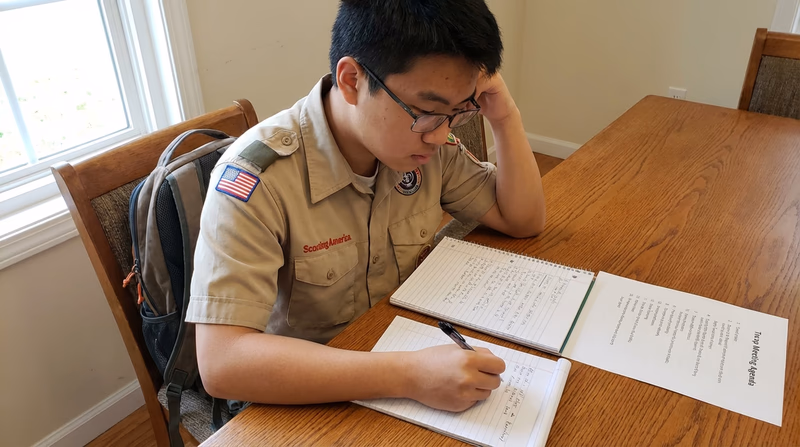 A Scout sitting at a desk reviewing their notes and writing a report, with a printed meeting agenda visible beside the notebook