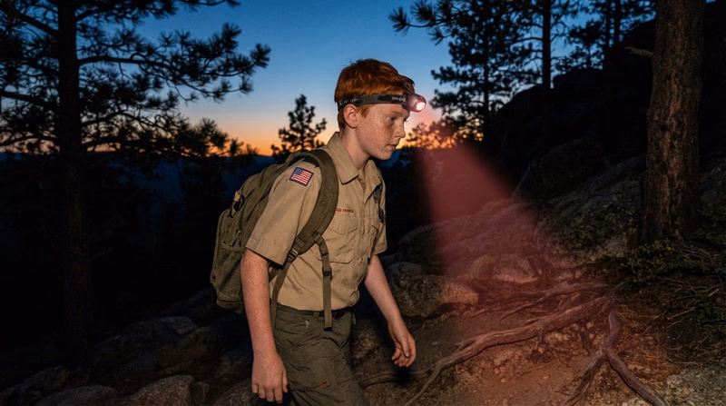 A Scout hiking at dusk with a headlamp illuminating the trail ahead