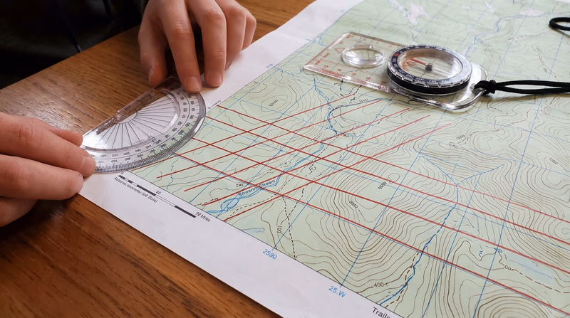 Hands drawing magnetic north-south lines on a USGS topographic map using a straightedge and red pencil, with a protractor at the starting point and several parallel red lines already drawn at a slight angle from the printed grid lines