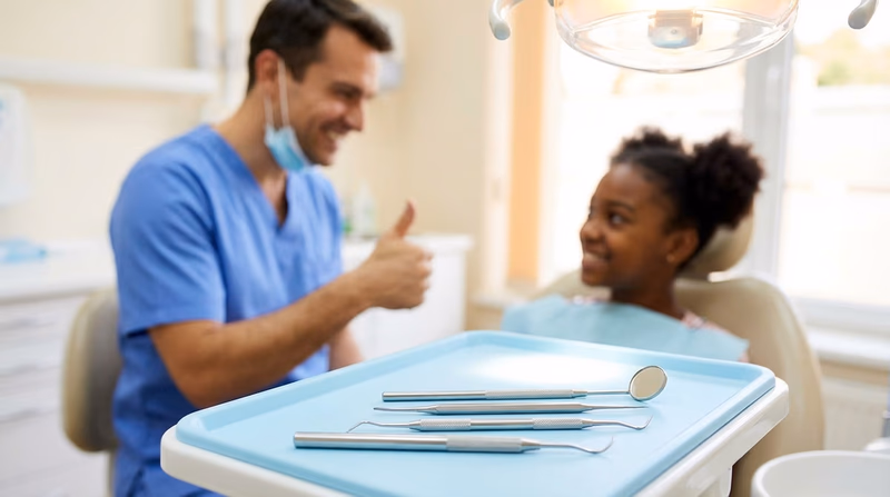 A close-up of dental tools arranged on a clean tray next to a dental chair, with a friendly dentist visible in the background