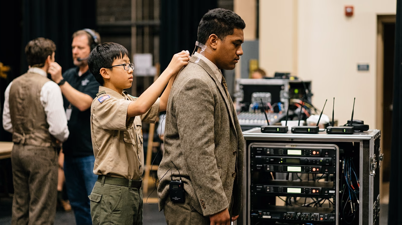 A sound technician backstage helping an actor adjust a wireless microphone pack attached to their costume, both looking focused