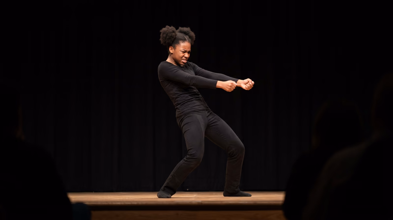 A teenager performing mime on a simple black stage, creating the illusion of pulling a rope, with focused expression and exaggerated body position